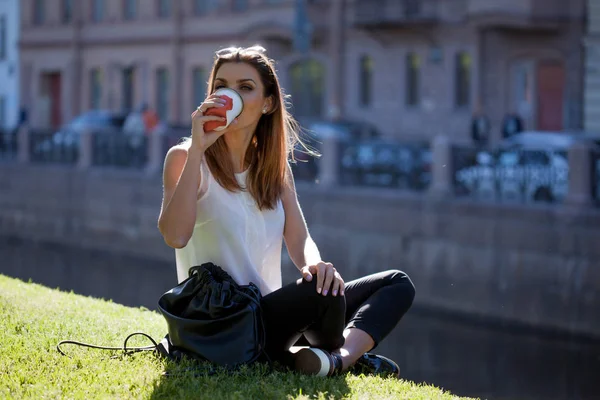 Молодая красивая женщина сидит на траве и пить кофе, чтобы пойти — стоковое фото