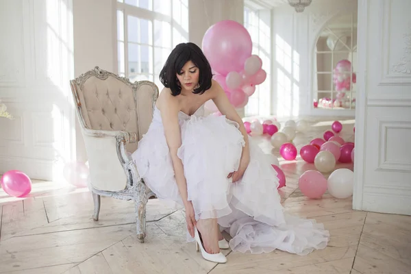 La novia se sienta en una silla y zapatos. Mujer joven en vestido de novia en el interior de lujo con una masa de globos de color rosa y blanco . —  Fotos de Stock