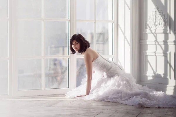 Retrato de joven morena atractiva con el pelo corto en un vestido de novia. Sentada cerca de la ventana, mirando a la cámara  . — Foto de Stock