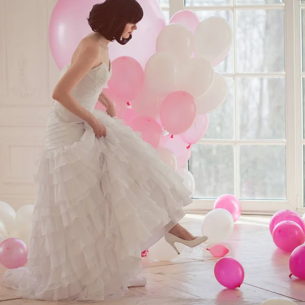 Mujer joven en vestido de novia en el interior de lujo con una masa de globos de color rosa y blanco. Sostenga en las manos sus zapatos blancos — Foto de Stock