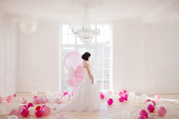 Young woman in wedding dress in luxury interior with a mass of pink and white balloons, standing against the window. — Stock Photo, Image