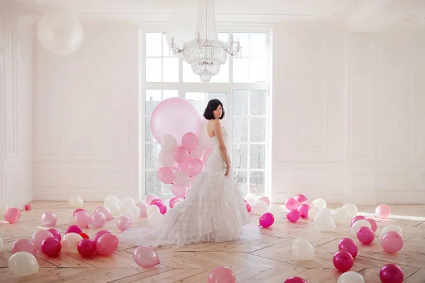 Mujer joven en vestido de novia en el interior de lujo con una masa de globos rosados y blancos, de pie contra la ventana . —  Fotos de Stock