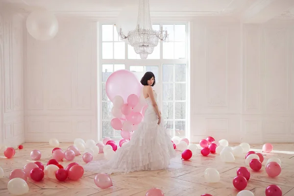 Young woman in wedding dress in luxury interior with a mass of pink and white balloons, standing against the window. — Stock Photo, Image