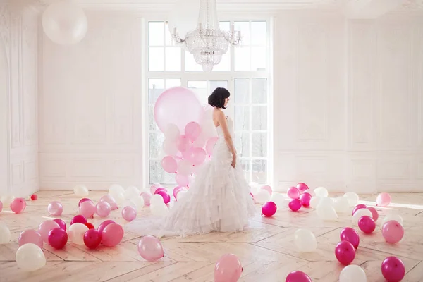 Mujer joven en vestido de novia en el interior de lujo con una masa de globos rosados y blancos, de pie contra la ventana . — Foto de Stock
