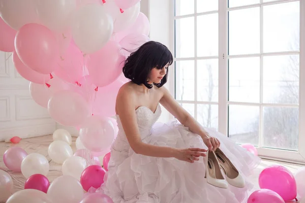 Young woman in wedding dress in luxury interior with a mass of pink and white balloons. Hold in hands her white shoes — Stock Photo, Image