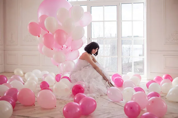 Jonge vrouw in trouwjurk in luxe interieur met een massa van roze en witte ballonnen. Haar witte schoenen in handen houden — Stockfoto