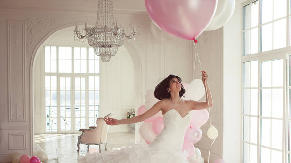 Young woman in wedding dress in luxury interior flies on pink and white balloons.