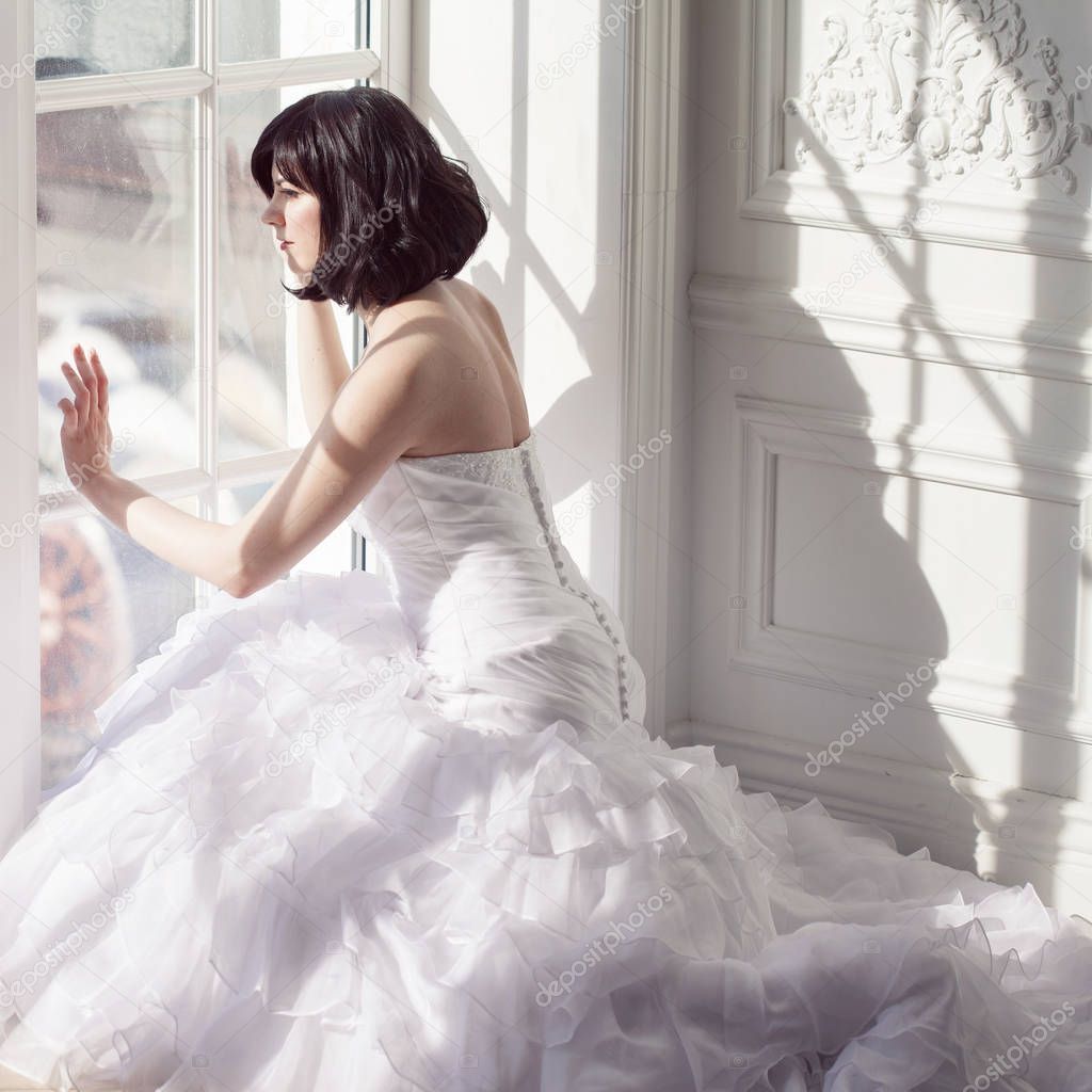 Portrait of young attractive brunette with short hair in a wedding dress. Sitting near the window, back view.