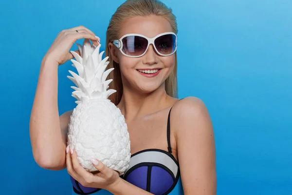 Mladá blond žena v modrých bikinách na modrém pozadí, sluneční brýle a bílé ananas — Stock fotografie