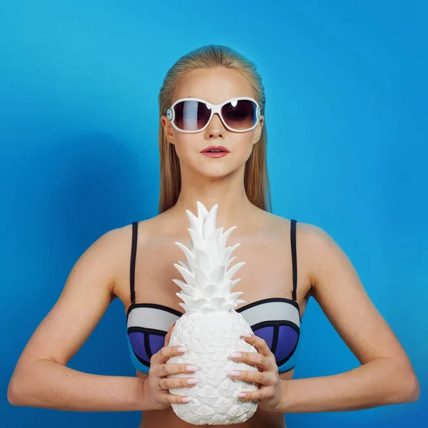 Молода блондинка в блакитному бікіні на синьому фоні, з сонцезахисними окулярами та білим ананасом — стокове фото