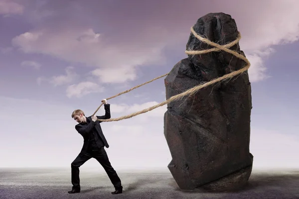 Businessman and a heavy burden, work concept. Young man in a suit is trying to move a huge stone — Stock Photo, Image