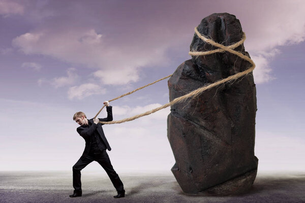 Businessman and a heavy burden, work concept. Young man in a suit is trying to move a huge stone