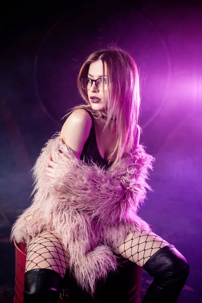 Jovem mulher atraente com roupas elegantes. Menina bonita no casaco de pele rosa fofo senta-se no barril. Luz de néon — Fotografia de Stock