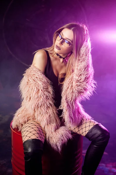 Young attractive woman with stylish clothes. Beautiful girl in fluffy pink fur coat sits on barrel. Neon light — Stock Photo, Image