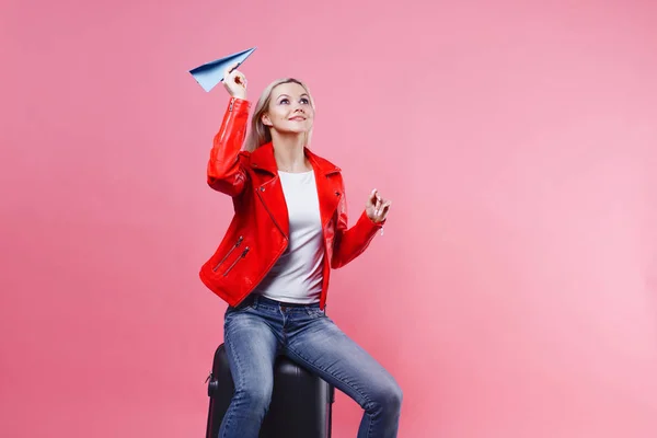 Счастливая молодая женщина с чемоданом для путешествий запускает бумажный самолет. Блондинка на розовом фоне, концепция — стоковое фото