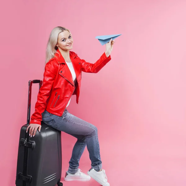 Glad ung kvinna med resor resväska lanserar pappersflygplan. Blond turist flicka på rosa bakgrund, begrepp — Stockfoto
