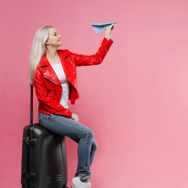 Glad ung kvinna med resor resväska lanserar pappersflygplan. Blond turist flicka på rosa bakgrund, begrepp — Stockfoto