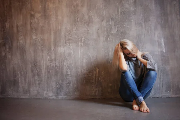 Triste mujer se sienta en el suelo. Depresión y fatiga crónica. Joven hermosa rubia en un suéter gris y jeans —  Fotos de Stock