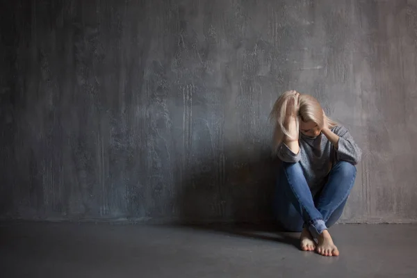 Triste mujer se sienta en el suelo. Depresión y fatiga crónica. Joven hermosa rubia en un suéter gris y jeans — Foto de Stock