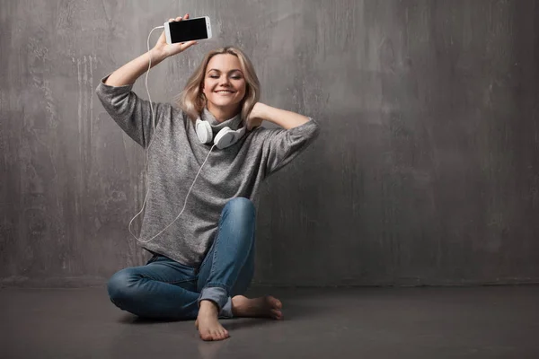 Νέοι και ελκυστική γυναίκα με smartphone και μεγάλες ακουστικά, κάθεται στο πάτωμα. Μουσική στην εφαρμογή για κινητά. — Φωτογραφία Αρχείου