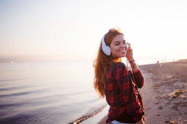 Happy attractive red-haired girl is enjoying music on walk. Beautiful young woman in shirt uses headphones, soft focus clipart