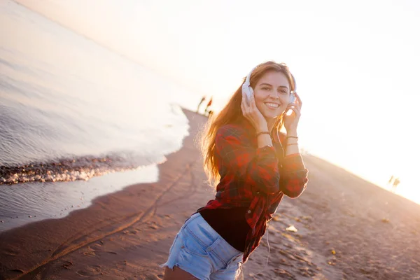 Happy attractive red-haired girl is enjoying favorite music on walk. Beautiful young woman in shirt uses headphones — Stock Photo, Image