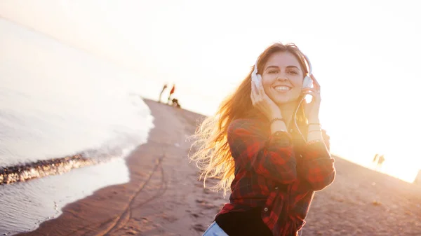 Счастливая привлекательная рыжеволосая девушка наслаждается любимой музыкой на прогулке. Красивая молодая женщина в рубашке использует наушники — стоковое фото