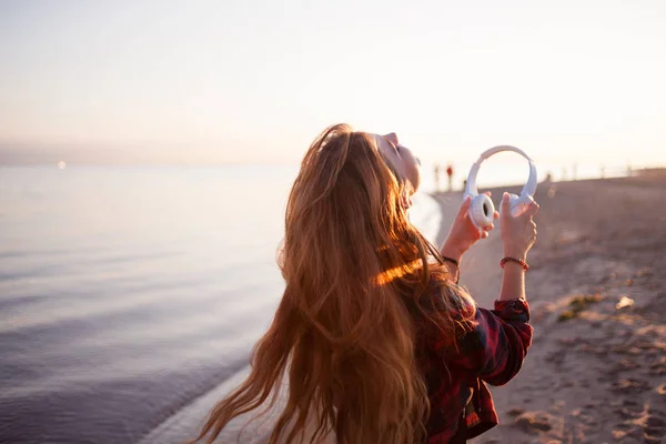 Happy attractive red-haired girl is enjoying music on walk. Beautiful young woman in shirt uses headphones, soft focus — Stock Photo, Image