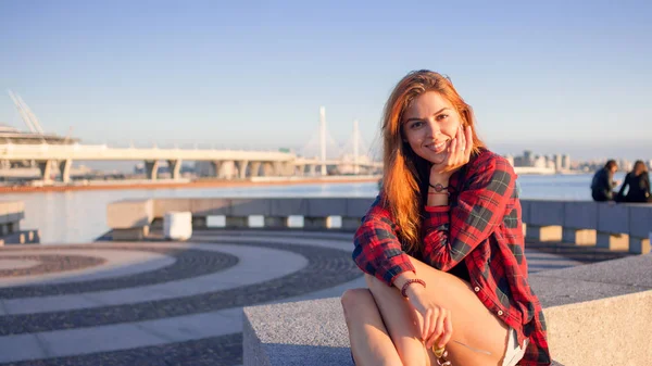 Linda jovem ruiva sentada à beira-mar, desfrutando da natureza e do bom tempo. A menina de camisa xadrez vermelho — Fotografia de Stock