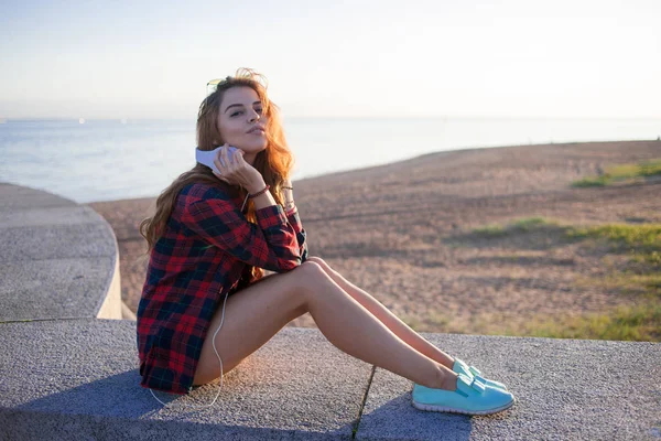 Piękna młoda Rudowłosa kobieta siedzi na nabrzeżu, zachwyt przyrodą i dobrej pogody. Dziewczyna w czerwonej koszuli w kratę — Zdjęcie stockowe