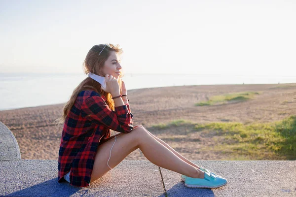 Hermosa joven pelirroja sentada en el paseo marítimo, disfrutando de la naturaleza y el buen tiempo. La chica de la camisa a cuadros roja — Foto de Stock