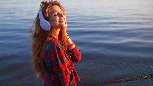 Happy attractive red-haired girl is enjoying favorite music on walk. Beautiful young woman in shirt uses headphones — Stock Photo, Image