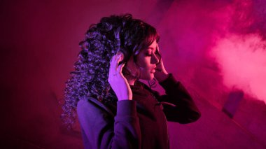 Büyük kulaklıklı genç, güzel, kıvırcık kadın müzik dinliyor, şarkı söylüyor, arka planda dans ediyor..