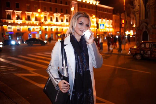Стильна молода жінка в елегантному бежевому пальто розмовляє по телефону, на міській вулиці вночі . — стокове фото