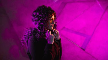 Büyük güneş gözlüklü genç, şık kıvırcık kadın, müziğe yöneliyor, kulaklıklarını çıkarıyor, dans ediyor, duvara yaslanıyor..