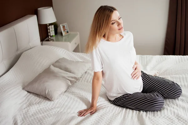 Mujer feliz embarazada calma está sentado en una cama ligera con almohadas, piernas cruzadas, descansando, pensando . — Foto de Stock