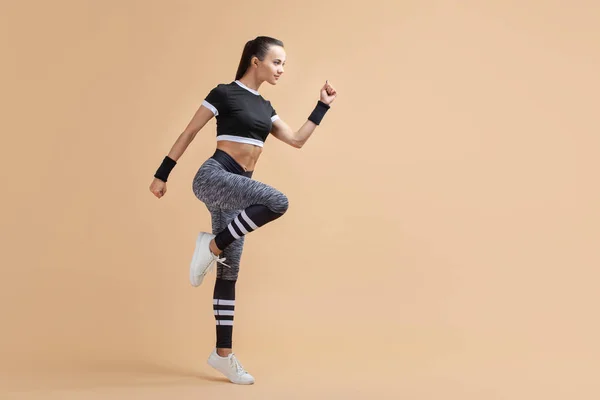 A young attractive brunette girl runs with wireless headphones raising her knees high on a peach background, cardio. — Stock Photo, Image