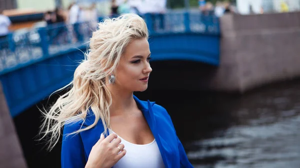 Retrato de una hermosa joven, una rubia con una chaqueta azul en el fondo del viejo puente azul sobre el río . —  Fotos de Stock