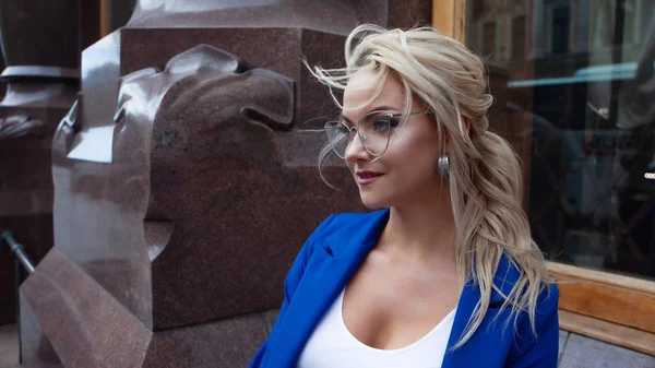 Portret pięknej blondynki w niebieskiej kurtce i stylowych okularach obok granitowego bollardu oraz szklane okno. — Zdjęcie stockowe