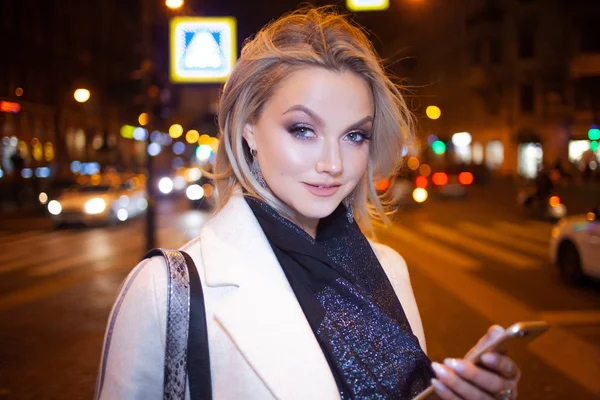 Стильна молода жінка в елегантному бежевому пальто використовує смартфон, що стоїть біля дороги, на вулиці міста вночі . — стокове фото