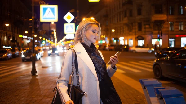 Zarif bej ceketli şık bir genç kadın, gece yarısı şehrin caddesinde duran bir akıllı telefon kullanıyor.. — Stok fotoğraf