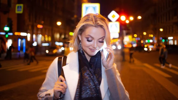 Stylowa młoda kobieta w eleganckim beżowym płaszczu używa smartfona stojącego przy drodze, na ulicy miasta w nocy. — Zdjęcie stockowe
