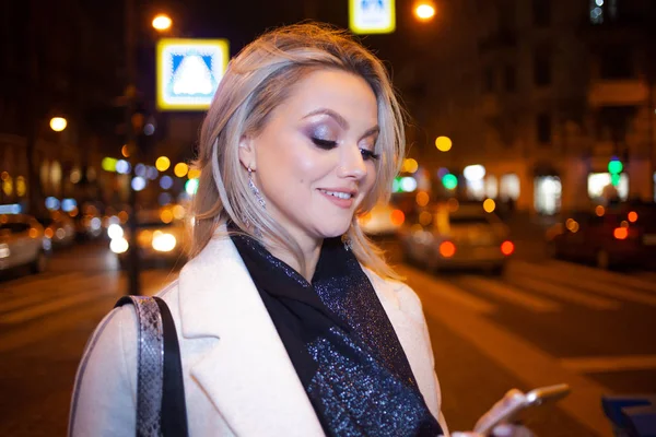 Snygg ung kvinna i en elegant beige rock använder en smartphone står vid vägen, på gatan i staden på natten. — Stockfoto