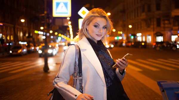 Snygg ung kvinna i en elegant beige rock använder en smartphone står vid vägen, på gatan i staden på natten. — Stockfoto