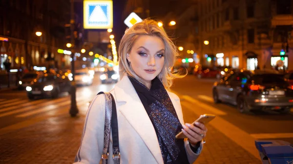 Mujer joven con estilo en un elegante abrigo beige utiliza un teléfono inteligente de pie junto a la carretera, en la calle de la ciudad por la noche . —  Fotos de Stock