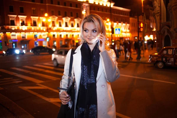 Stylische junge Frau im eleganten beigen Mantel nutzt ein Smartphone, das am Straßenrand steht, nachts auf der Straße der Stadt. — Stockfoto