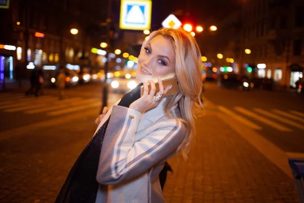 Stijlvolle jonge vrouw in een elegante beige jas maakt gebruik van een smartphone aan de weg, op de straat van de stad 's nachts. — Stockfoto