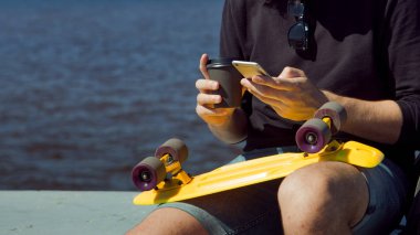 Kucağında sarı bir kaykayla rıhtımda oturan genç bir adam akıllı telefonu kontrol ediyor..