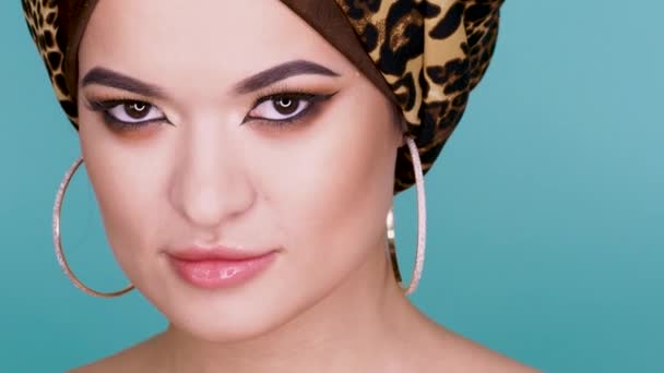Uma jovem atraente em um turbante elegante feito de tecido de impressão de leopardo em um fundo azul . — Vídeo de Stock