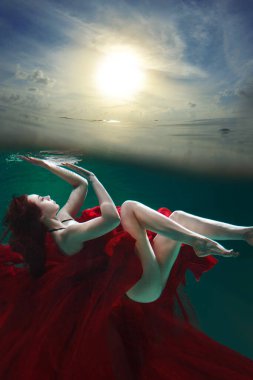 Mystical underwater portrait of a beautiful young woman in a red dress. The girl swims under the surface of the water